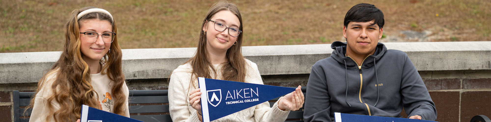 Three students sitting holding Aiken Technical College pennants