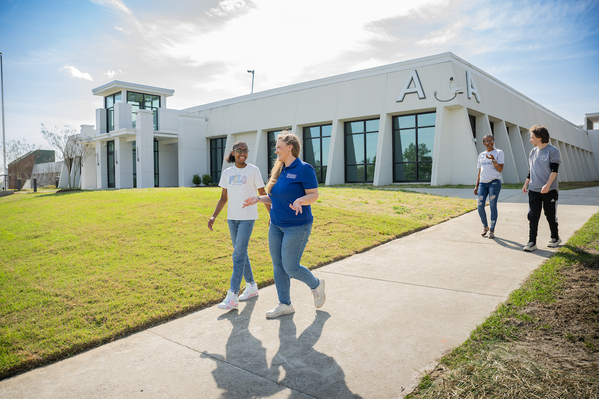 Four people walk along a sidewalk on a sunny day near a modern academic building with large windows, white exterior walls, and the letter “A” mounted on the front.