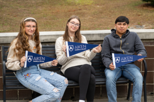 Three students sitting on an outside bench holding navy Aiken Technical College pennants