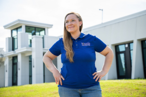 Alice Brown wearing a blue “Aiken Technical College” polo shirt standing outdoors in front of a modern campus building featuring large windows and white exterior walls, with a grassy lawn in the foreground