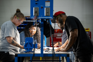 Three students work together at a table using a blue machine, with tools and equipment visible in a lab or workshop setting.